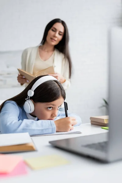 Babysitter floue avec livre près de la fille dans les écouteurs écriture pendant la leçon en ligne — Photo de stock