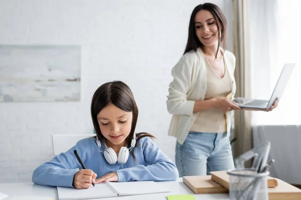 Smiling babysitter with laptop near girl writing in notebook at home — Stock Photo