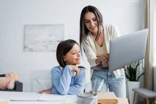 Babá mostrando laptop para menina sorridente fazendo lição de casa em primeiro plano desfocado — Fotografia de Stock