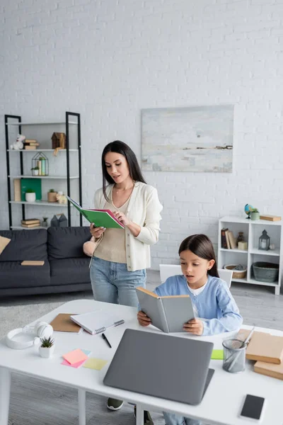 Child reading book while doing homework near laptop and babysitter — Stock Photo