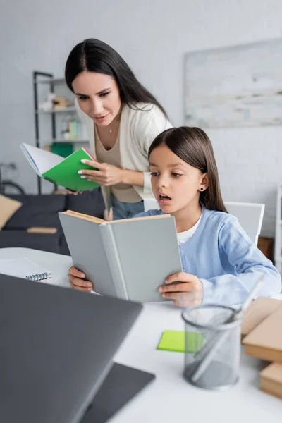 Babá com copybook perto de menina leitura livro enquanto fazendo lição de casa — Fotografia de Stock