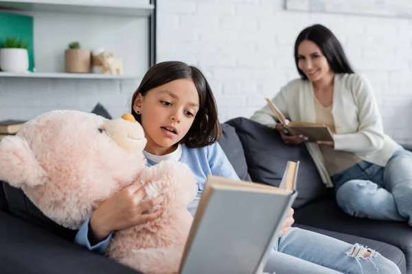 Fille embrassant ours en peluche tout en lisant le livre près de nounou souriant sur fond flou — Photo de stock