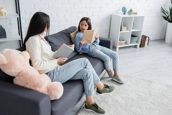 Longueur totale de l'enfant et nounou assis avec des livres sur le canapé dans le salon — Photo de stock