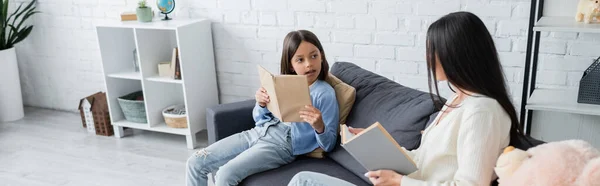 Girl looking at babysitter while reading book on couch in living room, banner — Photo de stock