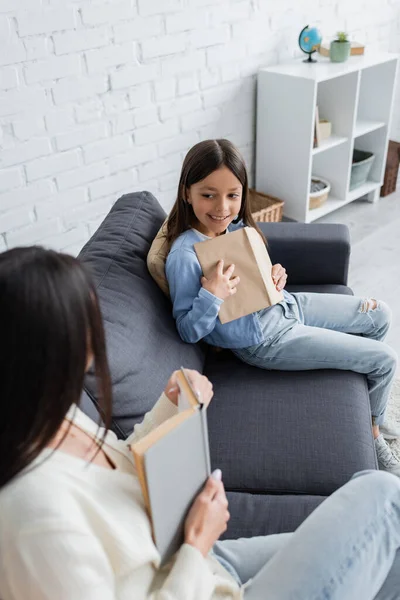 High angle view of smiling girl with book sitting on sofa near blurred nanny — Stock Photo