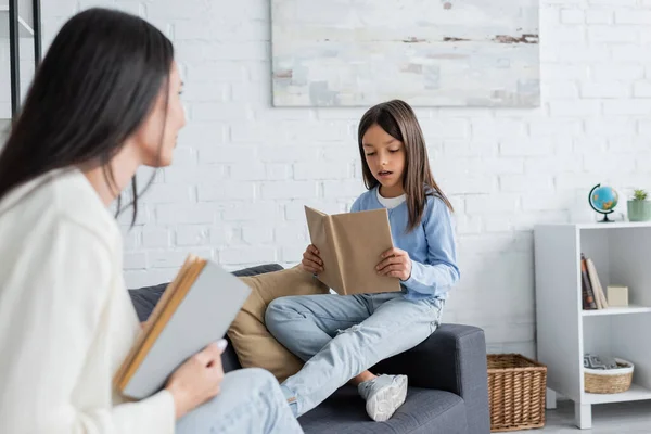 Ragazza seduta sul divano e lettura libro vicino babysitter offuscata — Foto stock