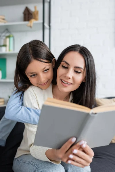 Lächelndes Mädchen umarmt Kindermädchen, das zu Hause Buch liest — Stockfoto