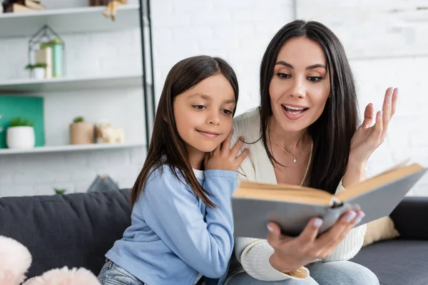 Babysitter lecture livre à sourire fille et geste sur flou au premier plan — Photo de stock