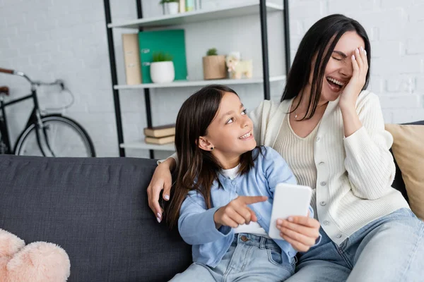 Laughing nanny obscuring face with hand near girl pointing at smartphone on couch at home - foto de stock