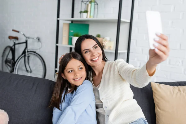 Smiling babysitter taking selfie on cellphone with girl on sofa at home — Foto stock