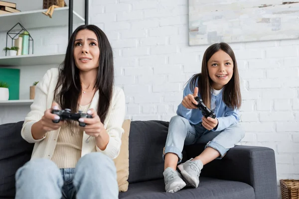 KYIV, UKRAINE - MAY 23, 2022: excited girl sticking out tongue while playing video game with worried nanny - foto de stock