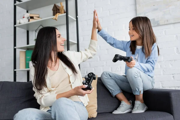 KYIV, UCRANIA - 23 DE MAYO DE 2022: niña feliz y niñera con joysticks dando cinco altos en el sofá en casa - foto de stock