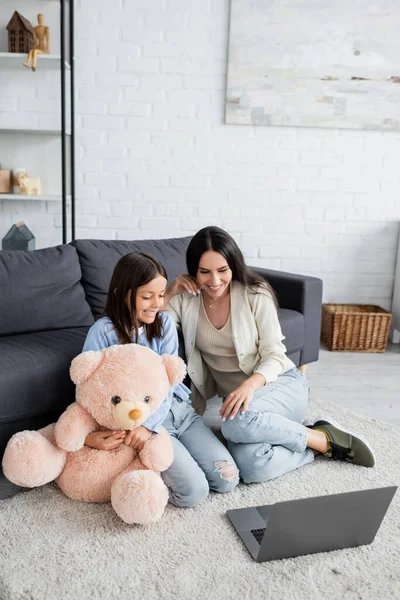 Happy girl and babysitter watching film on laptop while sitting on floor near teddy bear — Stock Photo