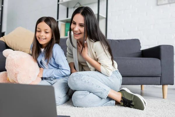 Alegre chica y niñera viendo la película en el ordenador portátil mientras está sentado en el suelo - foto de stock