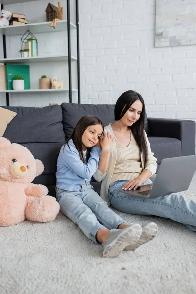Girl leaning on nanny while watching movie on laptop near teddy bear on floor — Fotografia de Stock