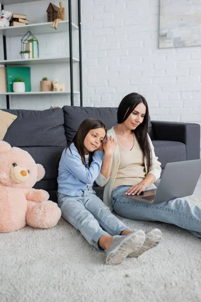 Fille penché sur nounou tout en regardant le film sur ordinateur portable près ours en peluche sur le sol — Photo de stock