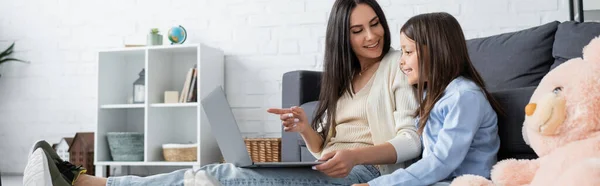 Babysitter pointing at laptop while watching film together with girl at home, banner — Foto stock