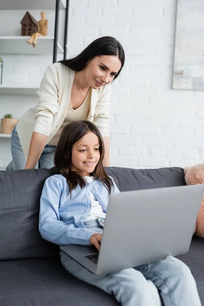 Baby-sitter souriant regardant fille jouer à un jeu vidéo sur ordinateur portable à la maison — Photo de stock