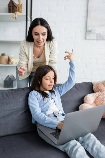 Excited girl gesturing with raised hand while gaming on laptop near nanny — Stock Photo