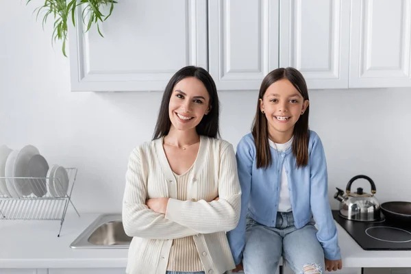 Happy girl sitting on kitchen counter near nanny standing with crossed arms — Stock Photo