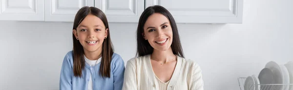 Cheerful babysitter and girl looking at camera in kitchen, banner — Stockfoto