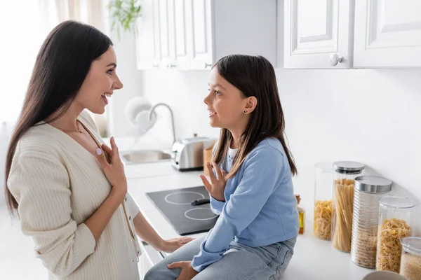 Smiling girl talking to excited nanny while sitting on kitchen counter near jars with pasta — Photo de stock