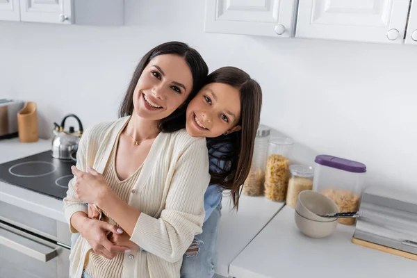 Glücklicher Babysitter mit lächelndem Kind, das sich in der Küche umarmt und in die Kamera blickt — Stockfoto
