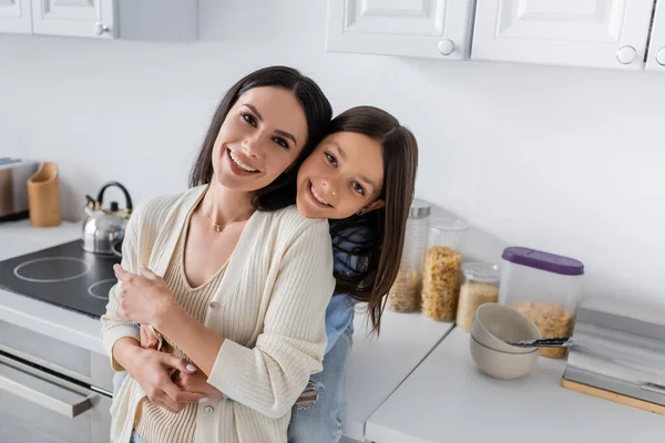 Babá feliz com criança sorridente abraçando e olhando para a câmera na cozinha — Fotografia de Stock