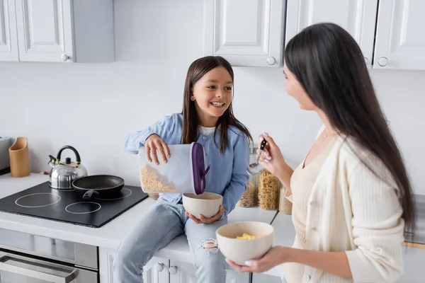 Feliz chica sentada en el mostrador de la cocina y verter copos de maíz en un tazón cerca de la niñera - foto de stock