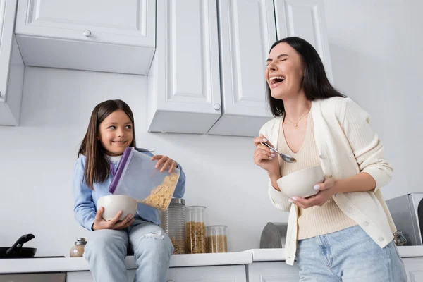 Lächelndes Mädchen, das Cornflakes ausschüttet, während es auf dem Küchentisch neben lachenden Kindermädchen sitzt — Stockfoto