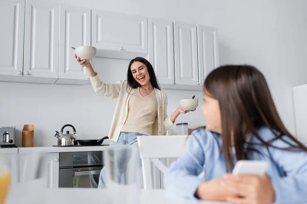 Збуджена няня тримає миски зі сніданком біля розмитої дівчини на кухні — стокове фото