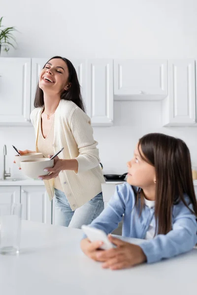 Baby-sitter excité tenant bols avec petit déjeuner et rire près de la fille dans la cuisine — Photo de stock