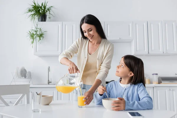 Cheerful babysitter pouring orange juice near child having breakfast in kitchen - foto de stock