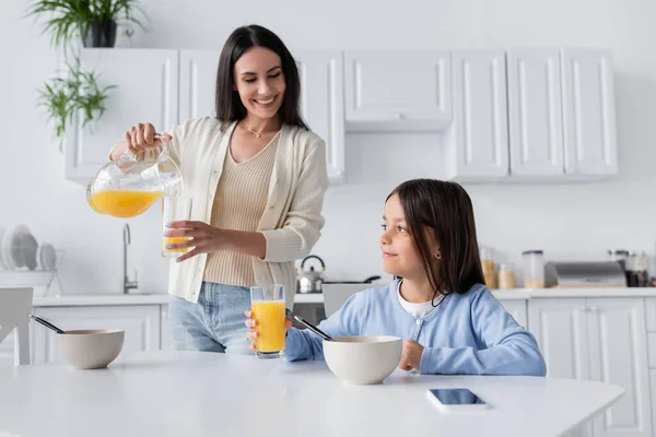 Bruna tata versando succo d'arancia vicino sorridente ragazza avendo colazione in cucina — Foto stock