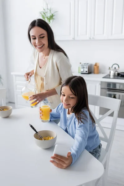 Smiling girl showing smartphone to nanny holding orange juice in kitchen - foto de stock