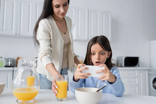 Изумленная девушка смотрит на мобильный телефон рядом с улыбающейся няней, подающей апельсиновый сок на завтрак — стоковое фото