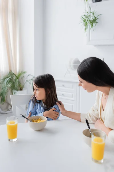 Nanny calming upset girl looking away near corn flakes and orange juice in kitchen — Stock Photo