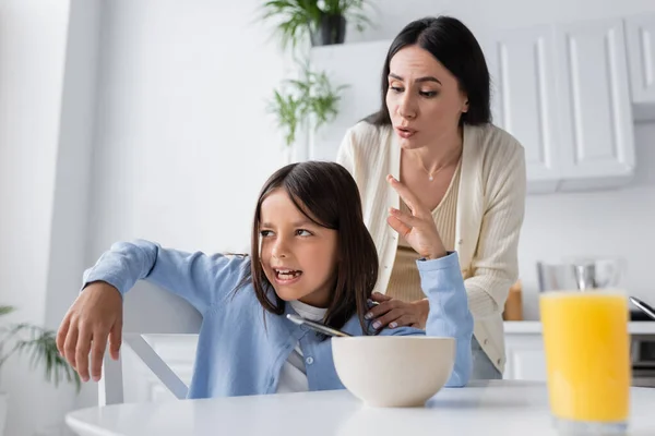 Irritated girl gesturing near breakfast and nanny calming her in kitchen — Stock Photo