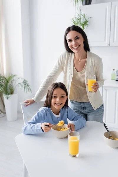 Menina feliz e babá olhando para a câmera perto de saborosos flocos de milho e suco de laranja fresco — Fotografia de Stock