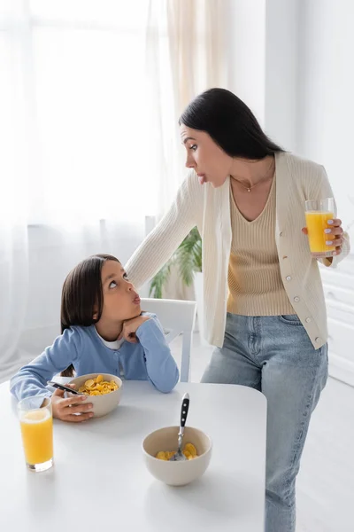 Nanny and girl looking at each other and sticking out tongues during breakfast in kitchen — Foto stock