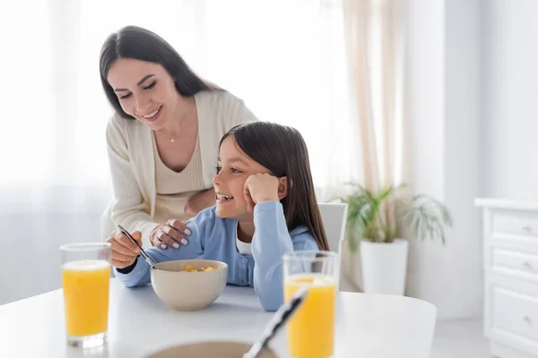 Happy babysitter looking at smiling girl having breakfast in kitchen — Stock Photo