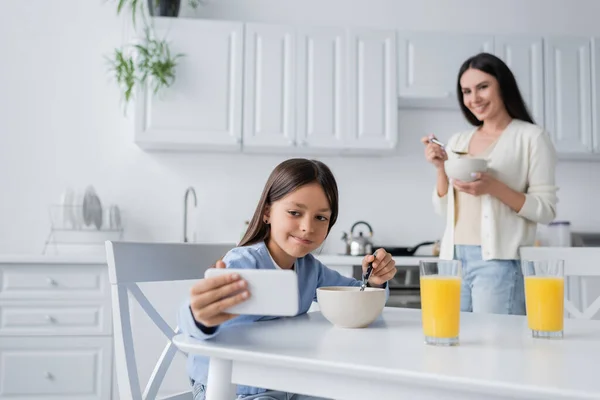 Gargalhada menina tomando selfie no smartphone durante o café da manhã enquanto babá sorrindo no fundo borrado — Fotografia de Stock