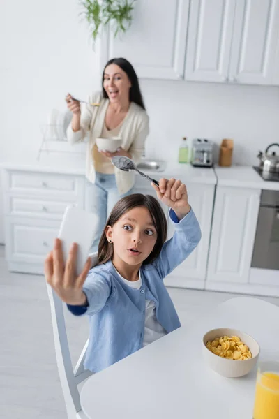 Excited girl with spoon taking selfie near corn flakes and cheerful nanny on blurred background - foto de stock