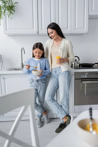 Piena lunghezza di ragazza mangiare corn flakes vicino bambinaia con succo d'arancia fresco — Foto stock