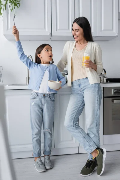 Chica asombrada sosteniendo cuchara en la mano levantada cerca de la niñera con un vaso de jugo de naranja - foto de stock