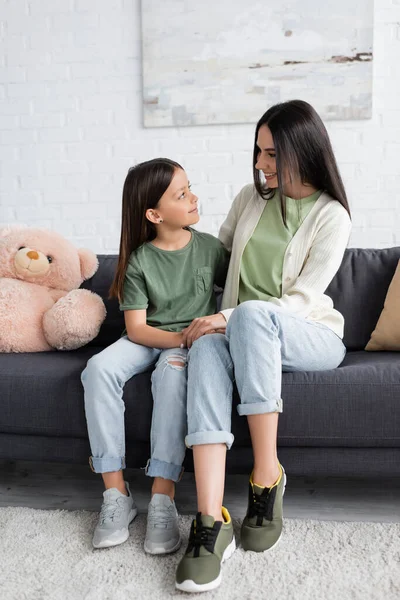 Full length of girl and nanny sitting on couch near teddy bear and smiling at each other — Stock Photo