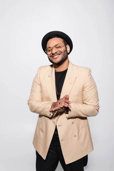 Happy indian hip hop performer in fedora hat and stylish eyeglasses smiling isolated on grey - foto de stock