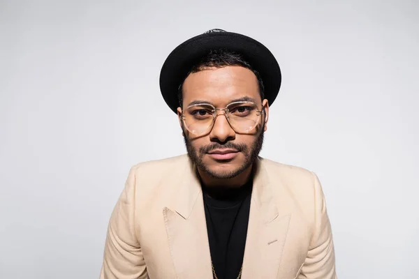 Stylish indian hip hop performer in fedora hat and eyeglasses looking at camera isolated on grey - foto de stock