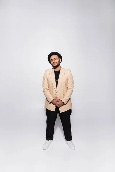 Full length of stylish indian hip hop performer in fedora hat and eyeglasses standing on grey — Stock Photo