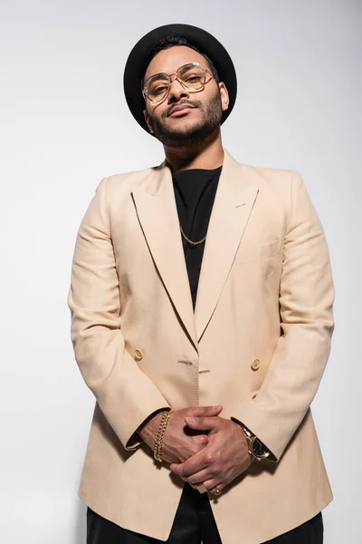 Low angle view of stylish indian hip hop performer in fedora hat and eyeglasses looking at camera isolated on grey — Fotografia de Stock